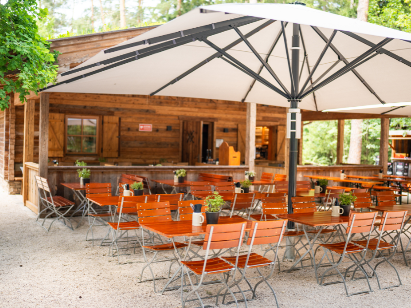 Fater Alm im Münchner Wald, Waldgarten mit Biertischen und - stühlen sowie Sonnenschirm als Überdachung.