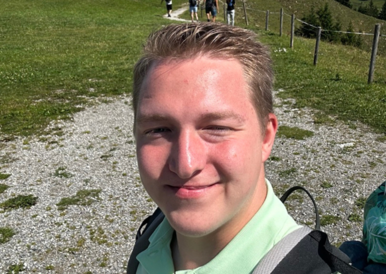 Daniel Demmel, Mitarbeiter der estermanngroup, lächelt in die Kamera. Er befindet sich auf einem Berg und trägt einen schwarzen Rucksack sowie Sportklamotten.