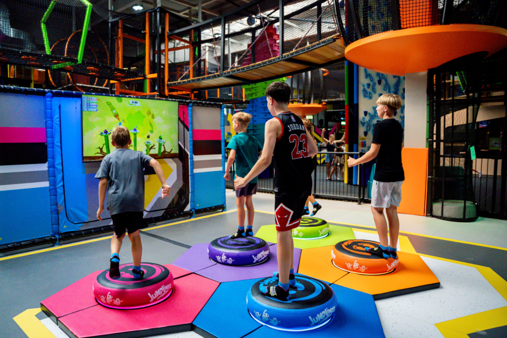 Fünf Jungs springen auf bunten Jumplings und spielen ein interaktives Videospiel. Vorne ist ein Bildschirm, auf welchem die Videospielfiguren Hinternisse meistern