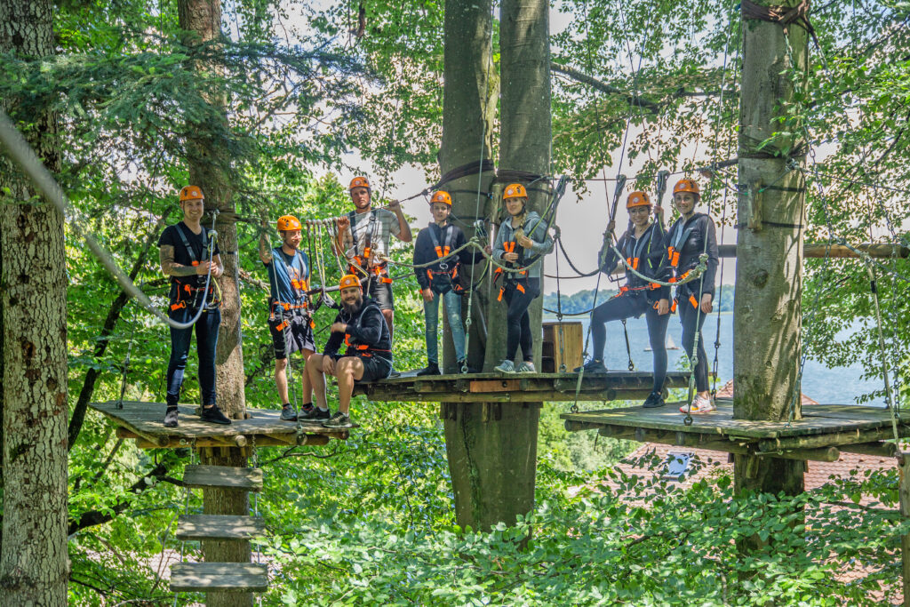Acht Erwachsene stehen gesichert auf Plattformen im Kletterwald Prien am Chiemsee. Sie lächeln in die Kamera und im Hintergrund sieht man die Kronen und Äste der Bäume sowie ein Blick auf den Chiemsee in Prien.