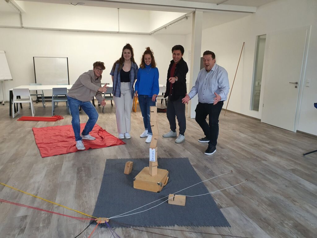 Sechs Personen stehen lachend um einen gemeinsam gebauten Holzturm in einem Seminarraum. Die Szene zeigt Teamarbeit, Kreativität und sichtbaren Stolz auf das Ergebnis.